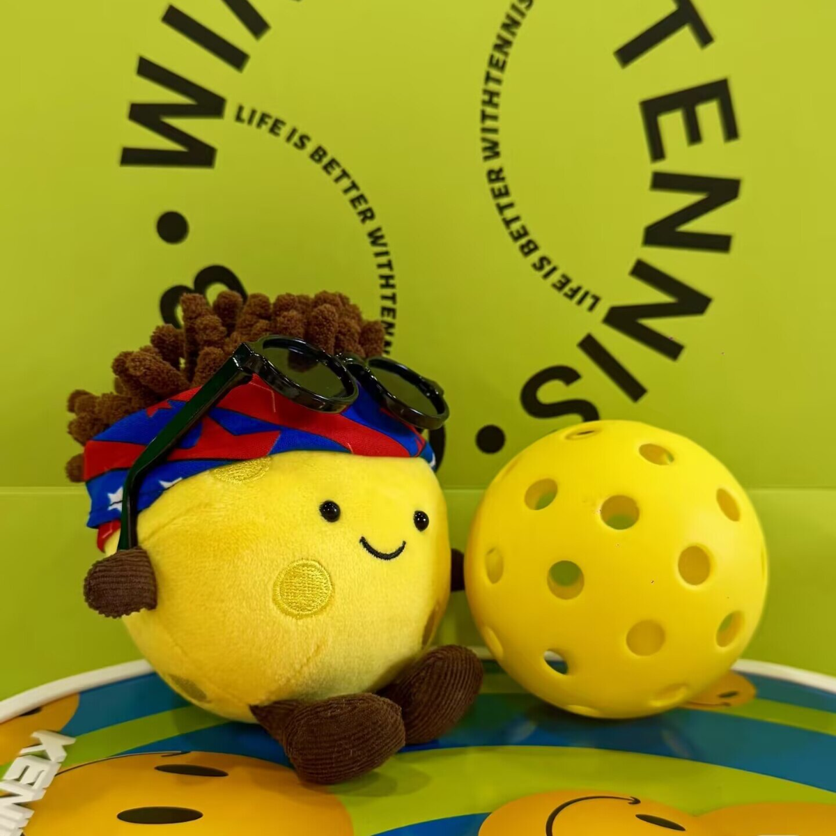 Plush pickleball character with sunglasses on a green background with tennis text