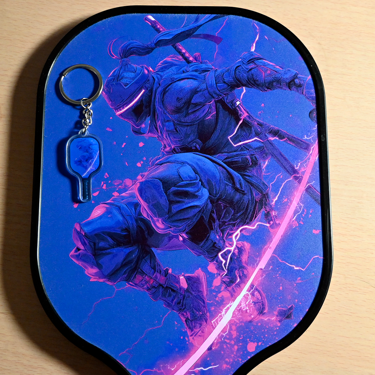 Keychain with a blue and pink graphic design of two warriors on a shield-shaped background.